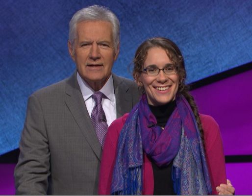 Grad student ashley omara stands next to jeopardy host alex trebek, she is wearing purple sweater and scarf and alex is wearing a suite and tie.