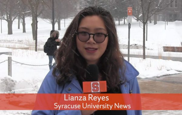 Lianza Reyes stands in front of Schine - she is holding a mic with orange mic flag, and there are students walking behind her in the background.