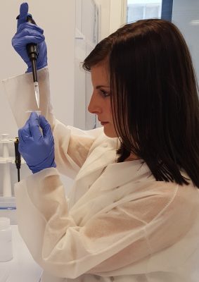 Kristina Gutchess, a Ph.D. candidate in Earth Sciences, works in a lab with a test tube. Gutchess is wearing a white lab coat and blue gloves.