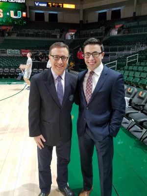 Ian Eagle stands next to his son Noah on the Miami basketball court. Both men are wearing suits and smiling. Both are wearing glasses
