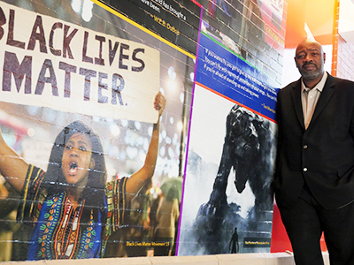 Professor Herb Ruffin standing to the right of a mural of Black-relevant art, such as a photo of a Black Lives Matter protester and a poster of Black Panter