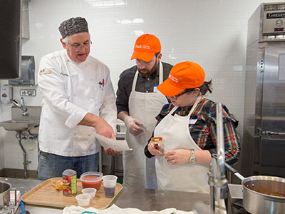 One man and two women work on preparing food; all wearing aprons and hats