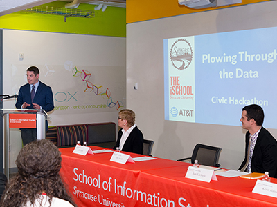 Syracuse Mayor Ben Walsh at podium on left, with iSchool Dean Liz Liddy seated in middle and City employee Sam Edelstein at right, with people in audience