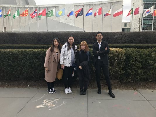 four students standing outside in front of row of international flags
