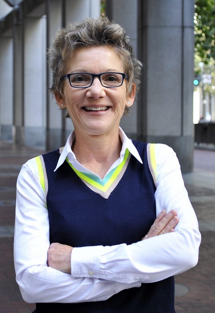 Person with short hair and glasses in a navy sweater vest standing with arms crossed in front of a columned building.