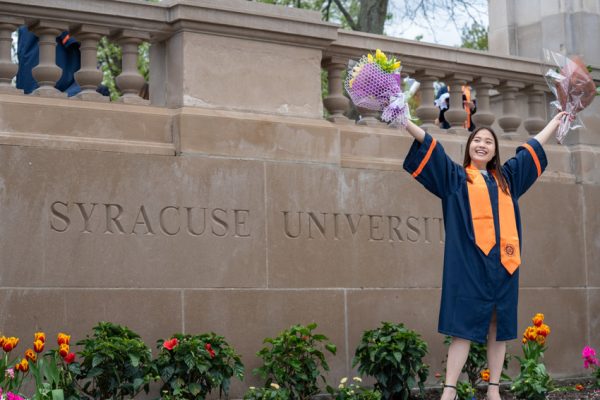 woman in graduation gown standing in front of Syracuse University sign etched in stone