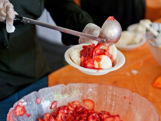person scooping strawberries over bowl of ice cream