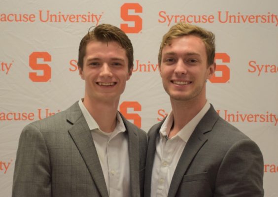 Two young men in suits in front of a Syracuse University banner