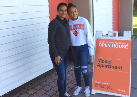 A mother and daughter stand outside the front door of an apartment. There is an open house sign signaling a model apartment next to them.