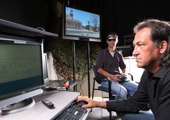Two men are in a room. A man sits in a chair wearing virtual reality gear while another man looks at a computer screen. 