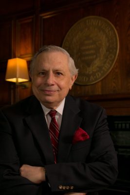 Person in a dark suit and red striped tie seated with arms crossed in front of a wooden wall and university emblem.