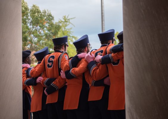 A group of men stand side-by-side with their arms on each other's backs. The photo is of their backs. They are wearing band uniforms with an "S" on the back. They are all wearing hats. Musical instruments are on the ground behind them.