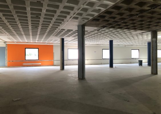 A large empty space in a building with 5 pillars and one orange wall. Five windows line the far wall. 