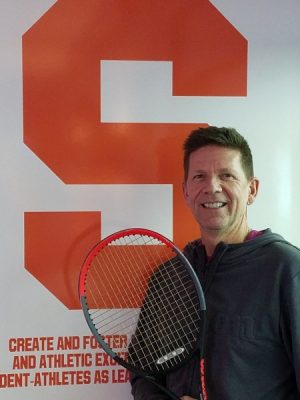 man posing with tennis racquet in front of Syracuse emblem