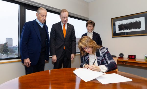 Three people standing watching woman sitting sign a document