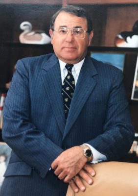 Person in a dark pinstripe suit standing in front of a leather chair and wooden shelves with framed photos and bird figurines.