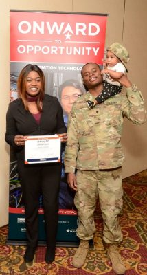 Person in a black suit holding a certificate stands beside a uniformed service member carrying a child, in front of an “Onward to Opportunity” banner.