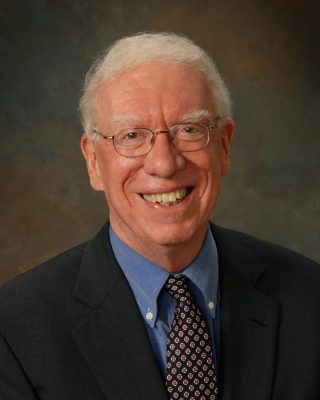 Person with white hair and glasses in a dark suit and patterned tie, smiling against a neutral brown background.