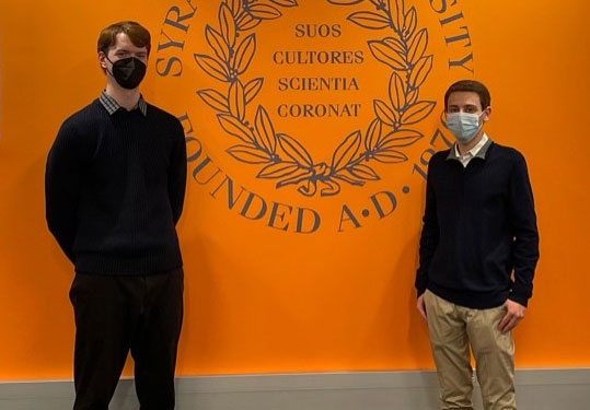 Ben Ayers and Zak Koeppel standing in front of wall with SU seal