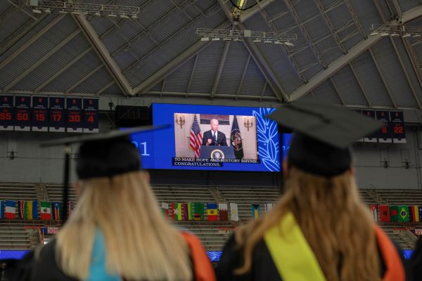 two students in caps and gowns watching a screen with President Joe Biden during Commencement 2021