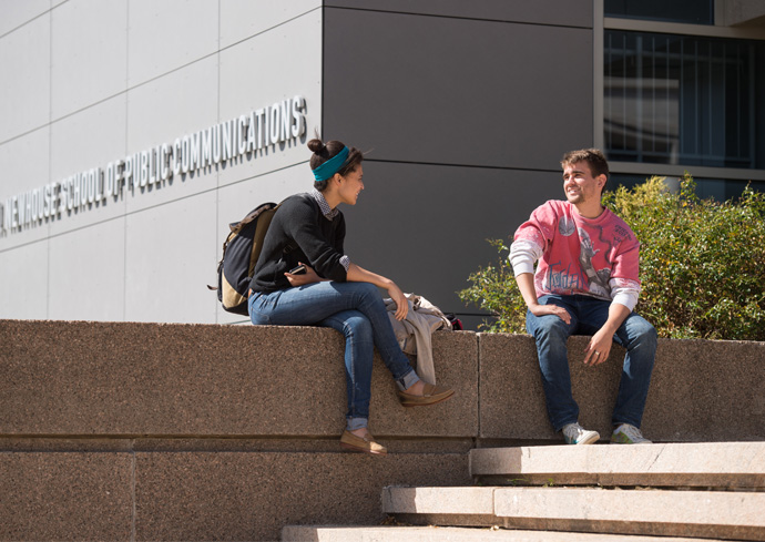 two students sitting on the granite wall outside of the Newhouse School, chatting amicably