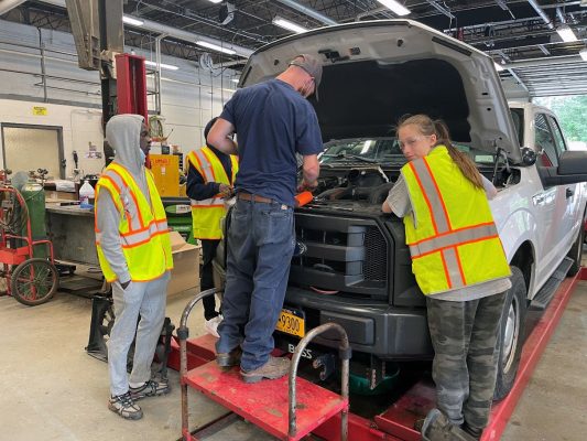 three people work beneath the hood of a motor vehicle