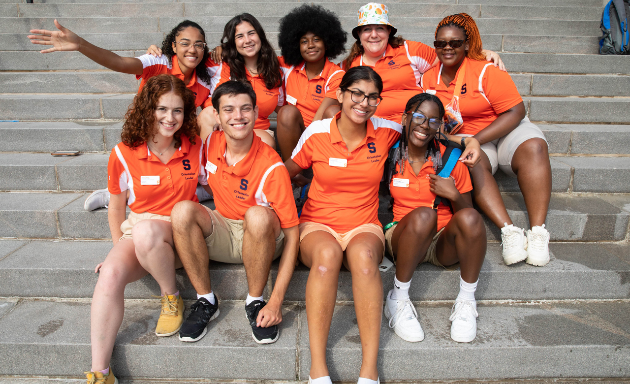 Nine orientation leaders in orange polo shirts huddle together for a picture on steps during Syracuse Welcome