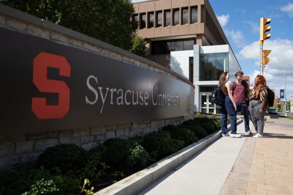 several students gather near the Syracuse University sign installed on the wall between Schine Student Center and the Newhouse complex