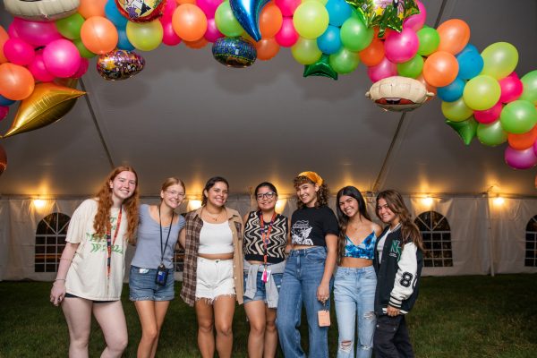 seven students gather together and smile under a colorful array of balloons at 90s night on the Quad