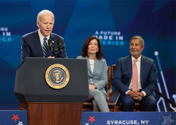President Biden speaks at a podium at Onondaga Community College on Oct. 27, with Gov. Kathy Hochul and Micron CEO Sanjay Mehrotra seated over his left shoulder