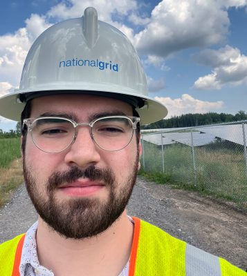 Selfie of Wyatt Bush wearing a yellow reflective vest and National Grid hardhat against a background of blue sky and clouds