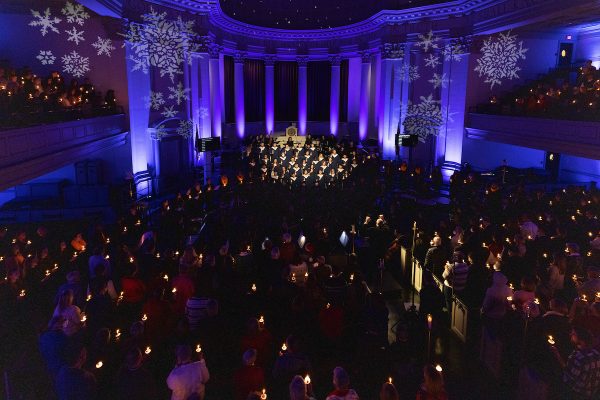 Room full of people in Hendricks Chapel holding candles with purple lighting and a chorus singing at the front facing the audience