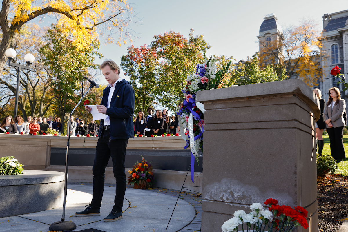 Young man standing at a microphone reading a piece of paper, standing outside on a sunny day at the Remembrance Wall