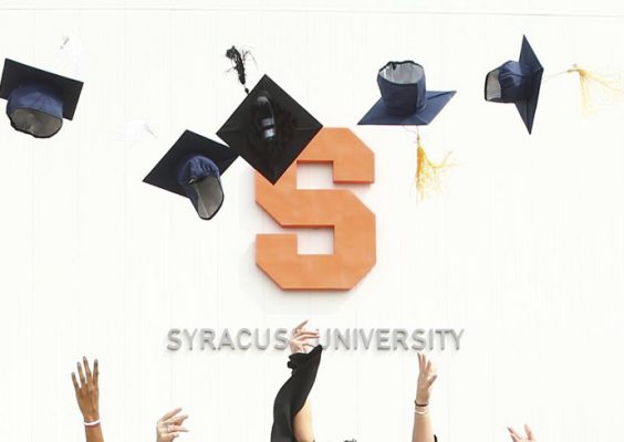 graduates throw caps in the air in front of a Syracuse University sign