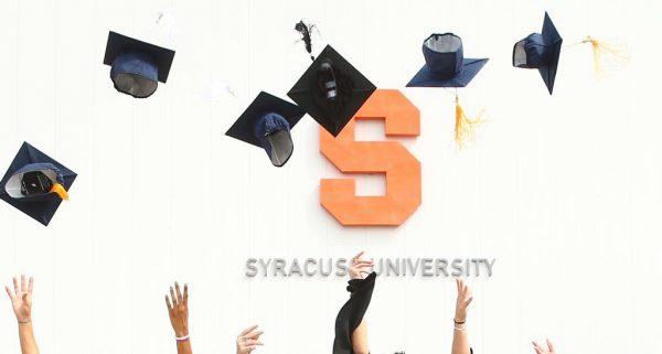 graduates throw caps in the air in front of a Syracuse University sign