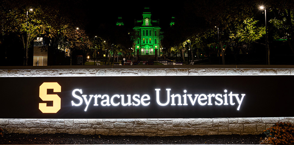 Hall of Languages lit up in green with a view of the Syracuse University sign