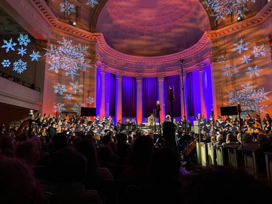 Hendricks Chapel lit up with orange and blue lighting while a chorus signs for an audience during Holidays at Hendricks.