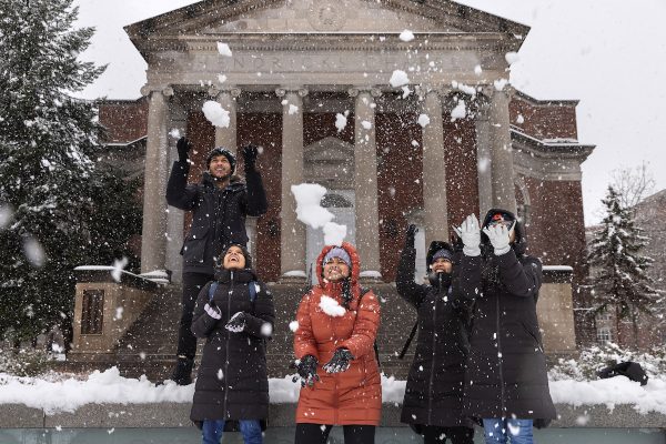 Several students standing outside throwing snowballs in the air while it is snowing heavily.