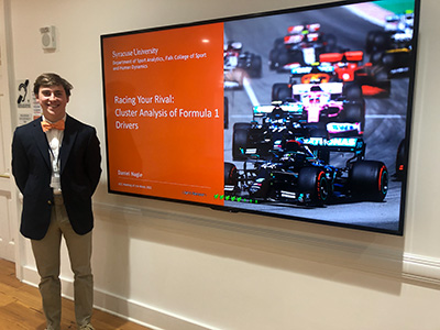 Young man standing next to a television with a PowerPoint slide on the screen with a race car on it. 