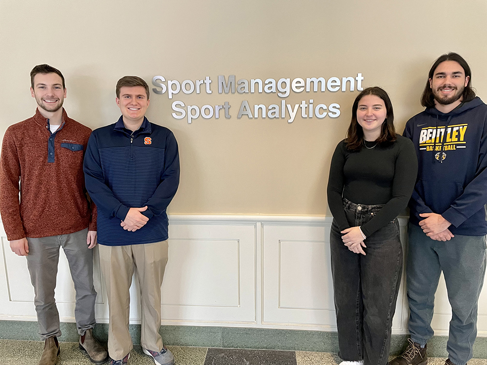 Students Eli Miller, Shane Halpin, Kylie Dedrick and Corey Goldman pose in front of lettering that says "Sport Management Sport Analytics" in the Falk College