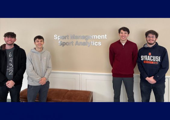 Students Matthew Gennaro, Alexander Borelli, Sam Gelman and Benjamin Wachtel pose in front of lettering that says "Sport Management Sport Analytics" in the Falk College