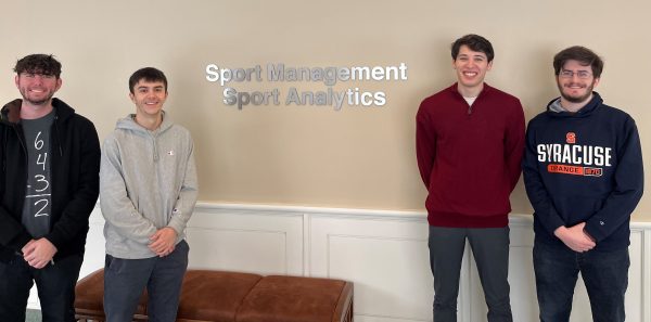Students Matthew Gennaro, Alexander Borelli, Sam Gelman and Benjamin Wachtel pose in front of lettering that says "Sport Management Sport Analytics" in the Falk College