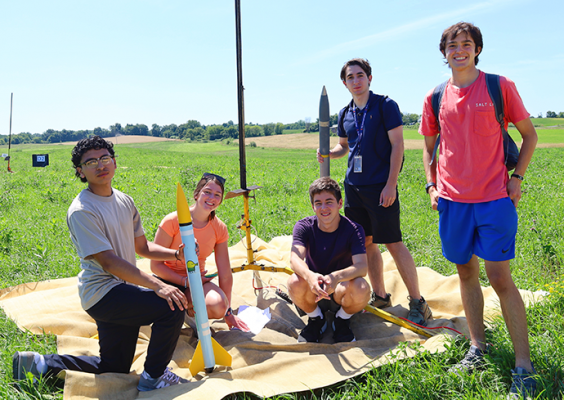 Five individuals outside on a sunny day working with small rockets