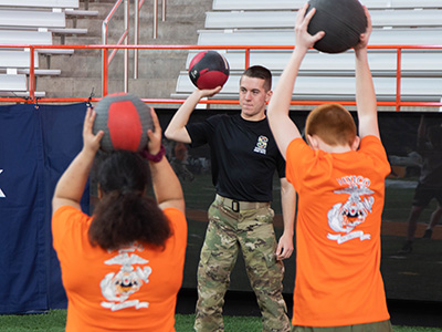 Two people in orange t-shirts hold medicine balls over their heads with an instructor in black facing the camera, doing the same