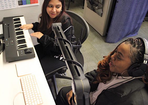 One individual sitting playing a keyboard sitting next to a person wearing headphones and talking into a microphone
