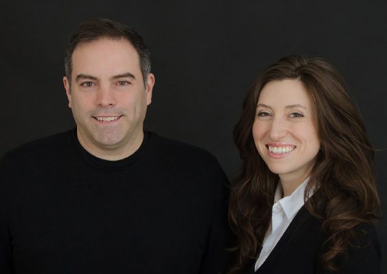 A man and a woman smile in front of a black backdrop.