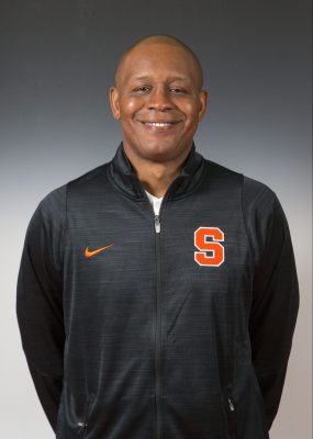 A man smiles for a headshot while wearing a blue Syracuse men's basketball warmup jacket.
