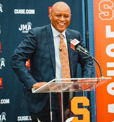 Man smiling while standing in front of a microphone with the Syracuse Athletics backdrop.