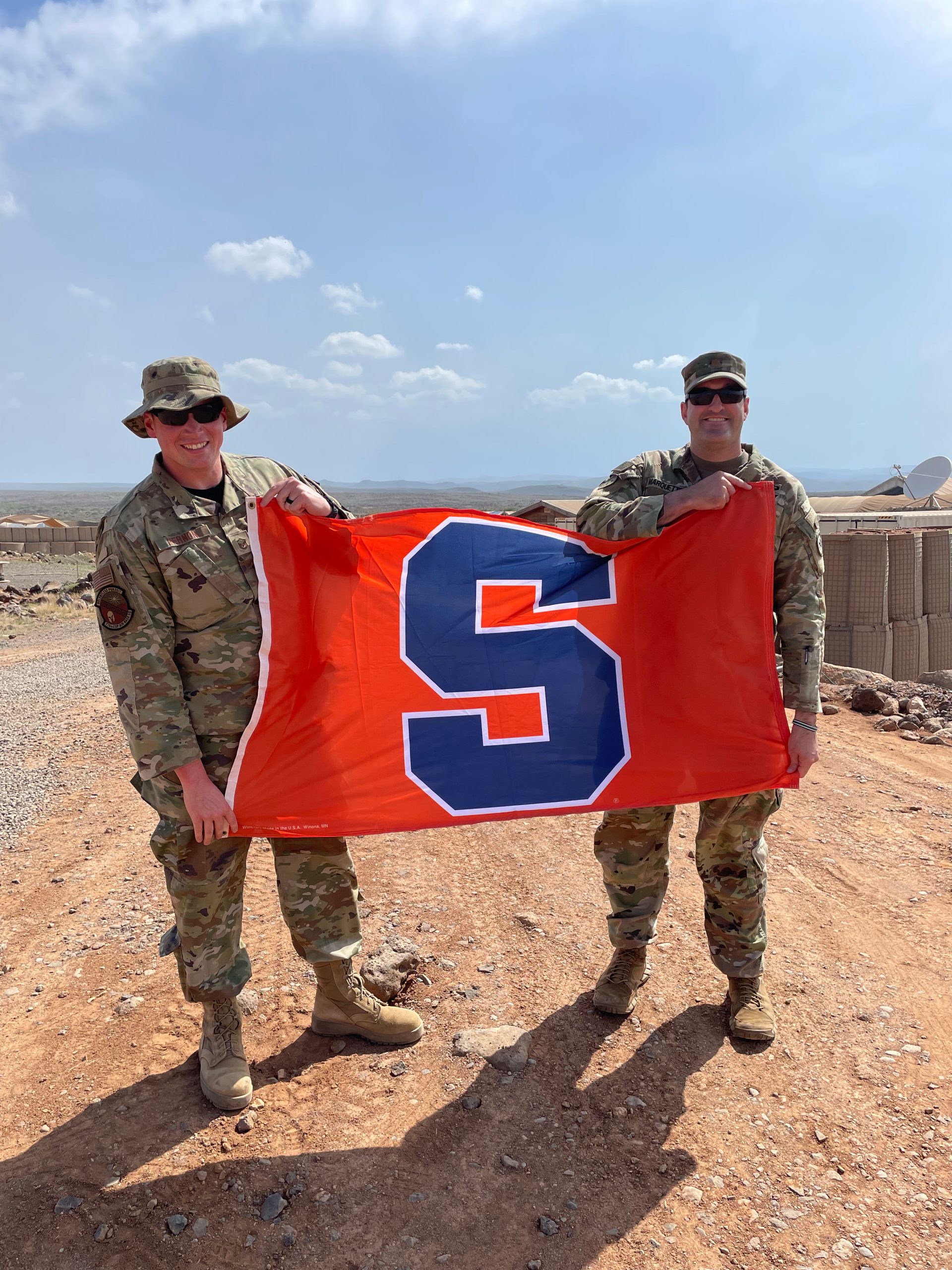 Two individuals in military uniform standing together holding a orange and blue Syracuse flag.