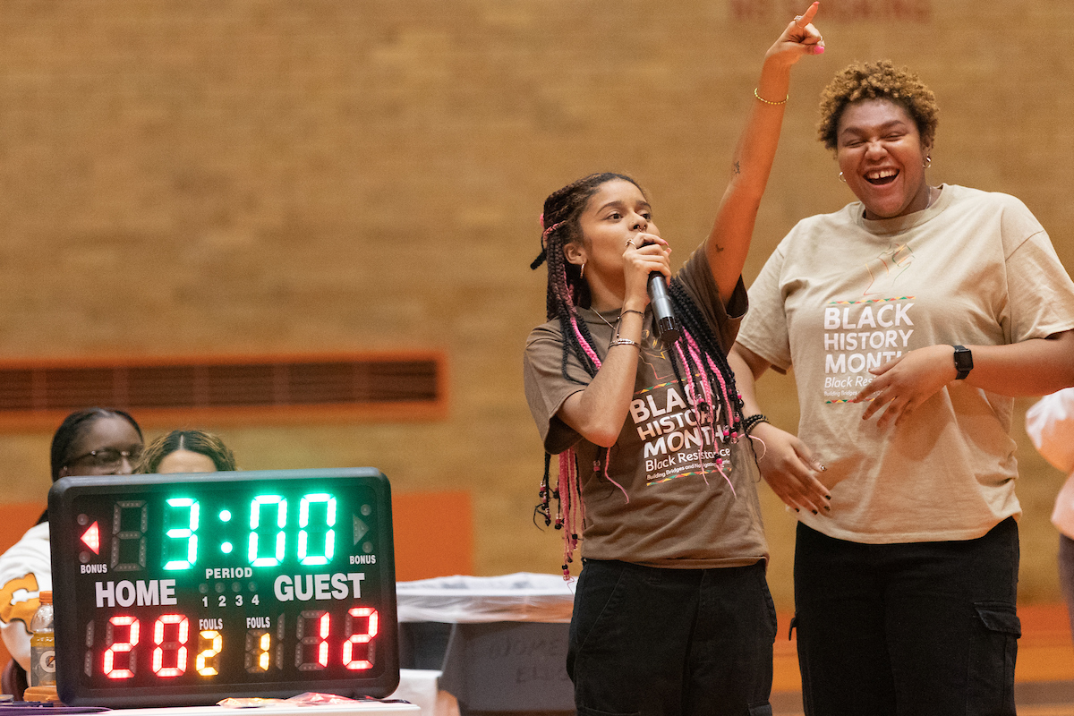 person speaking into a microphone next to a tabletop scoreboard during the annual basketball classic game